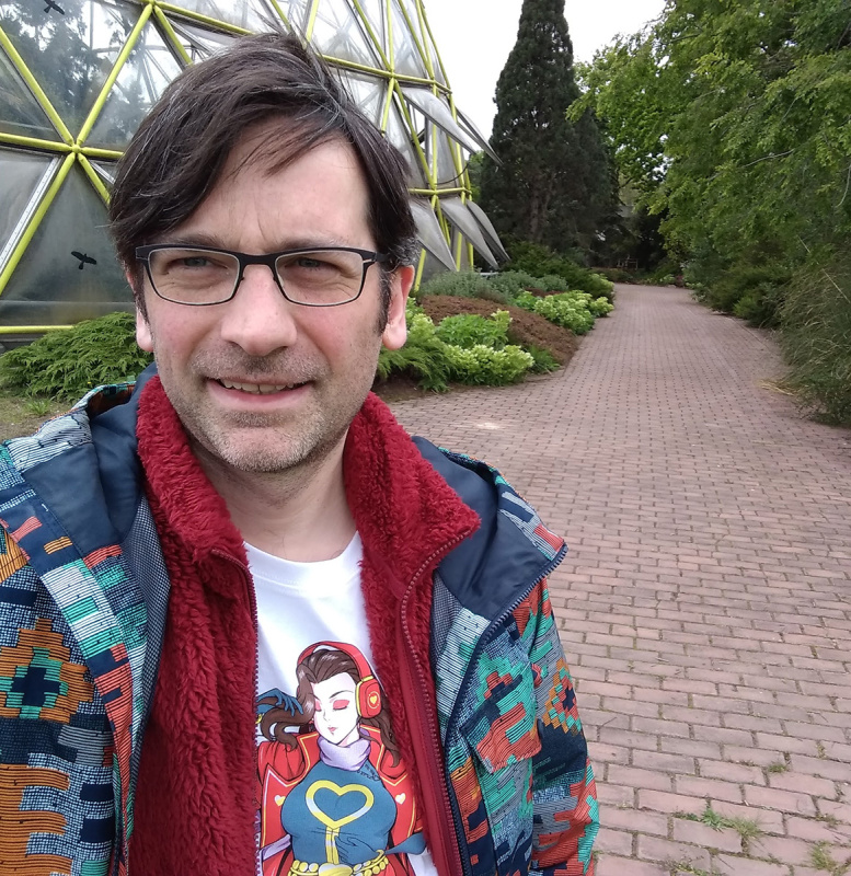 Selfie einer Person mit dunklen Haaren und Brille in einer parkähnlichen Anlage