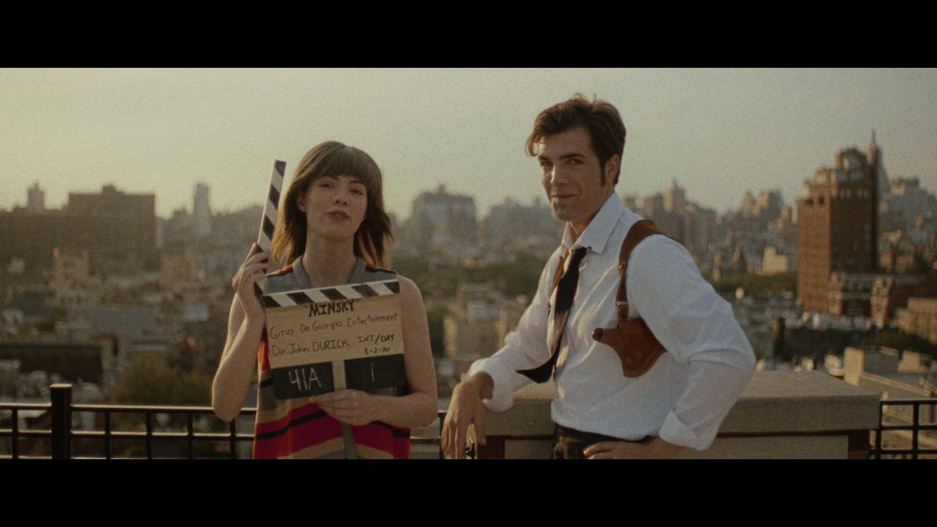 Eine Frau mit einer Filmklappe und ein Mann stehen auf dem Dach eines Hauses in New York, im Hintergrund Hochhäuser; das Bild ist mit einem Filter versehen, wodurch es wie altes Filmmaterial aus den 1970ern wirkt