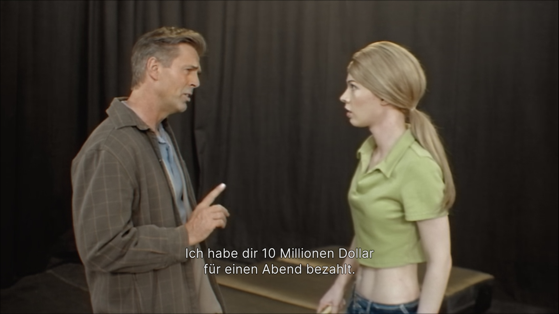Foto einer Probe für eine Filmszene; ein älterer Mann mit erhobenem Zeigefinger sagt zu einer sehr jungen, erschrockenen Frau: 'Ich habe die 10 Millionen Dollar für einen Abend bezahlt'
