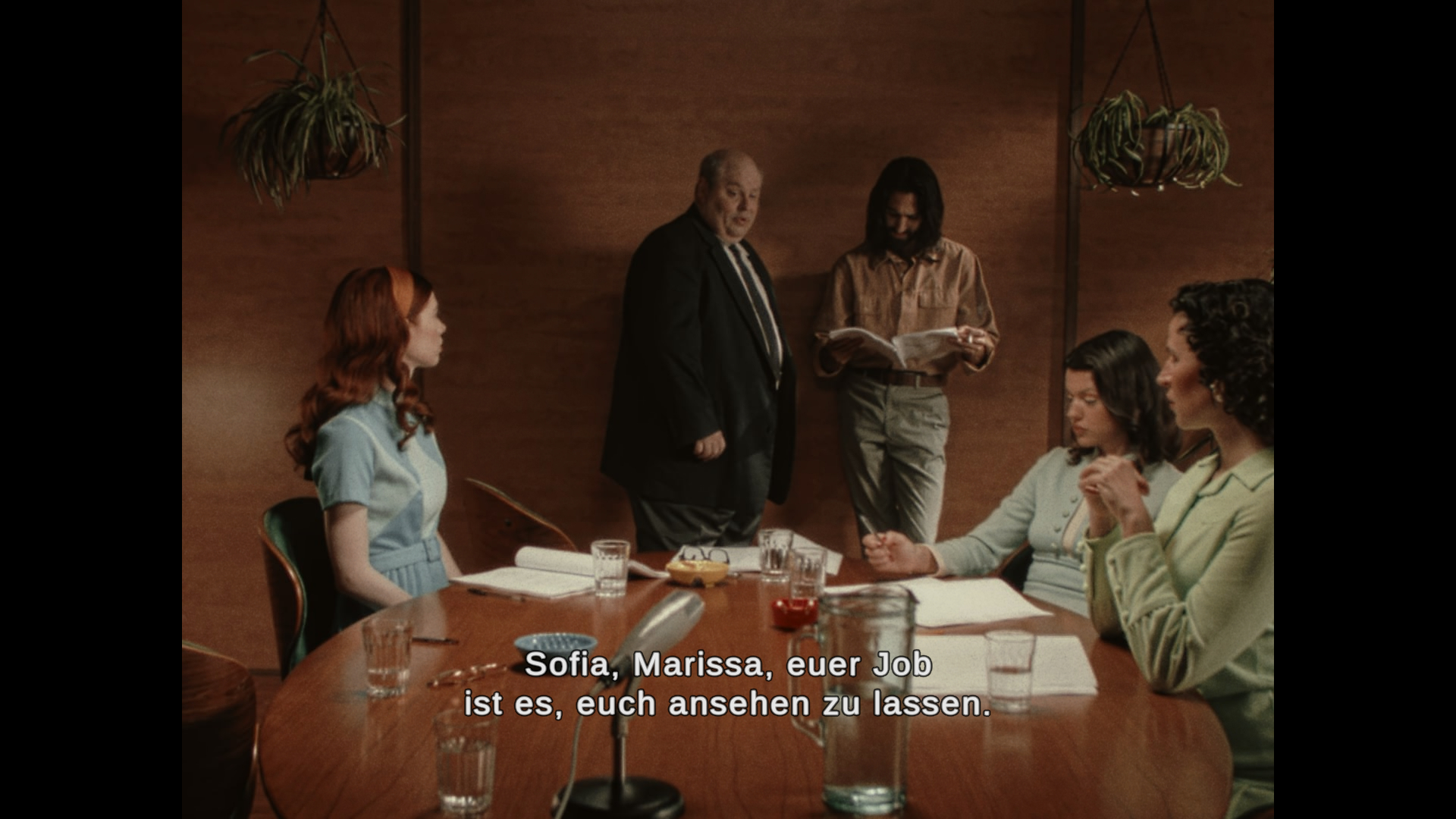 Foto eines Besprechungsraums; drei Frauen sitzen an einem Tisch, ein Mann steht an eine Wand gelehnt und liest in einem Skript; ein alter Regisseur im Anzug sagt zu zwei der Frauen: 'Sofia, Marissa, euer Job ist es, euch ansehen zu lassen'.