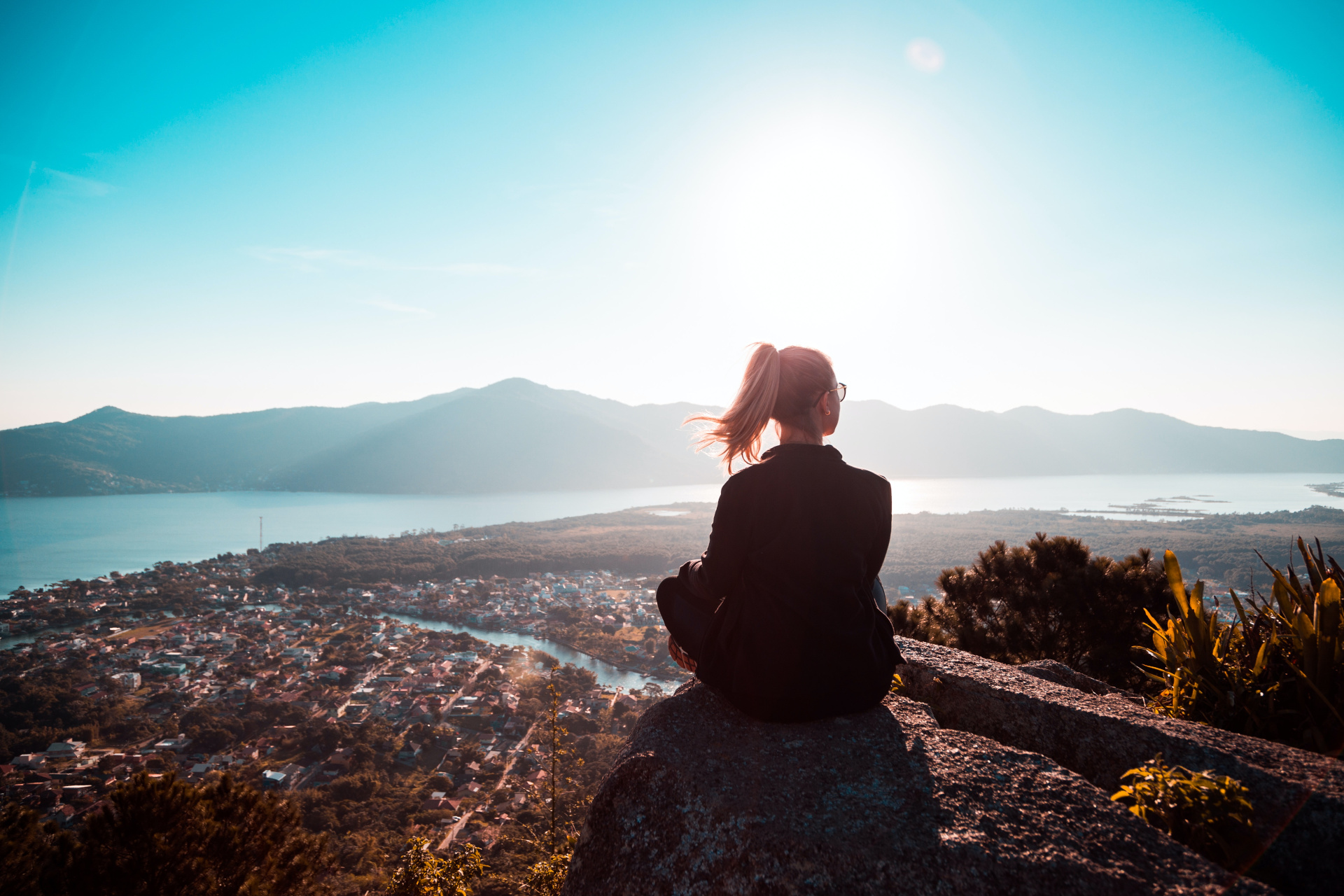 eine Frau sitzt in einer weiten Landschaft, der Himmel ist blau und sonnig, etwas weiter vor und unter ihr ist das Meer.
